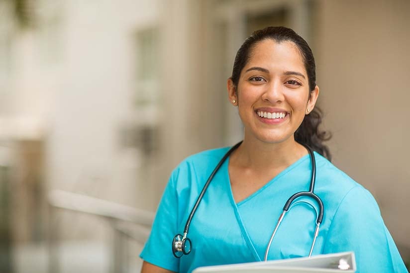A cheerful nurse in turquoise scrubs, with a stethoscope around her neck, holds a clipboard and smiles warmly, standing in a well-lit hospital corridor.